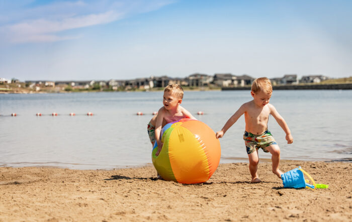 Copilot said: Two boys sharing a beach ball on the sandy beach at Harmony Lake, enjoying a sunny day by the water with the lake and shoreline in the background.