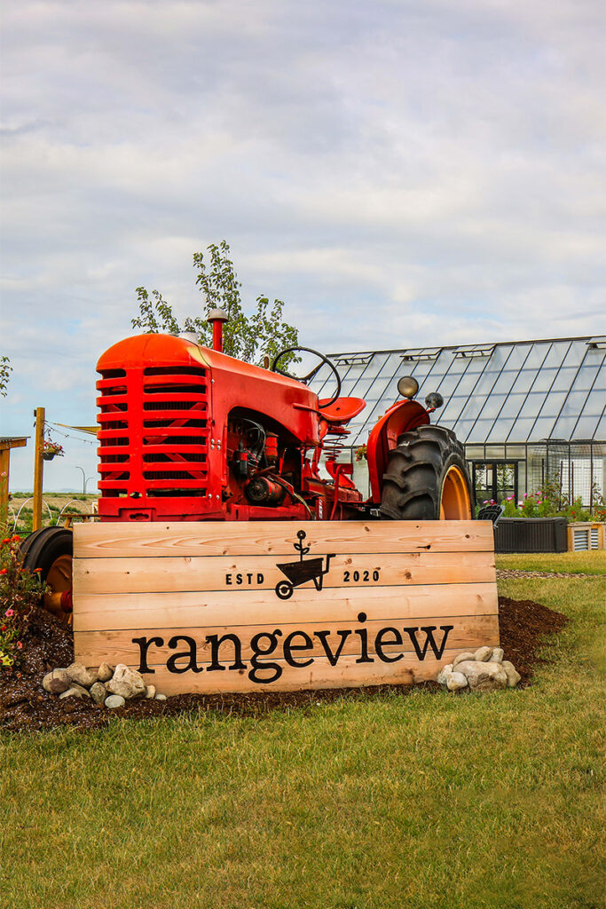 Rangeview community sign displayed beside a red tractor with the community greenhouse in the background, highlighting the garden-to-table lifestyle that makes Rangeview a unique Calgary community.